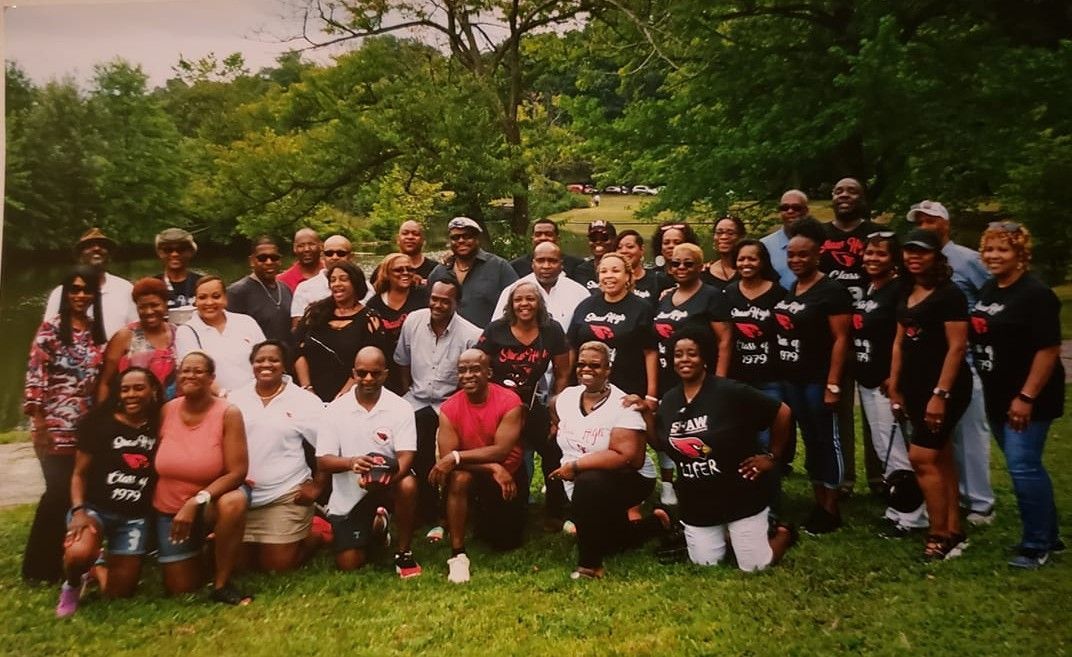 Large group of diverse people posing for a photo outdoors near trees and water. Some wear matching shirts.
