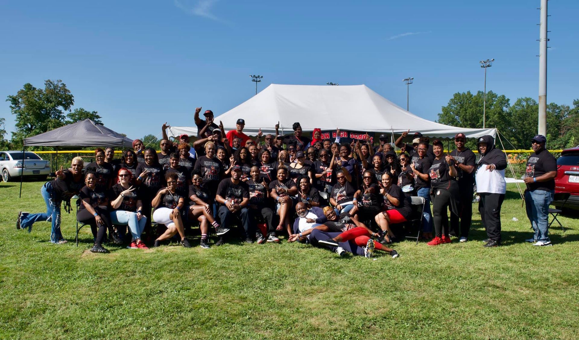 Large group of people in black shirts gather on a grassy field under a tent on a sunny day.