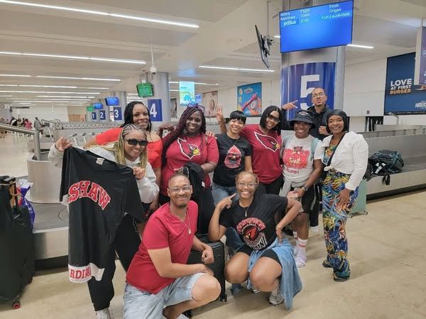 Group of diverse people smiling at an airport baggage claim, holding shirts and posing for a photo.