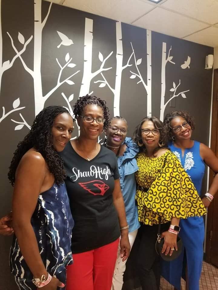 Five smiling women posing in front of a wall with white tree designs.