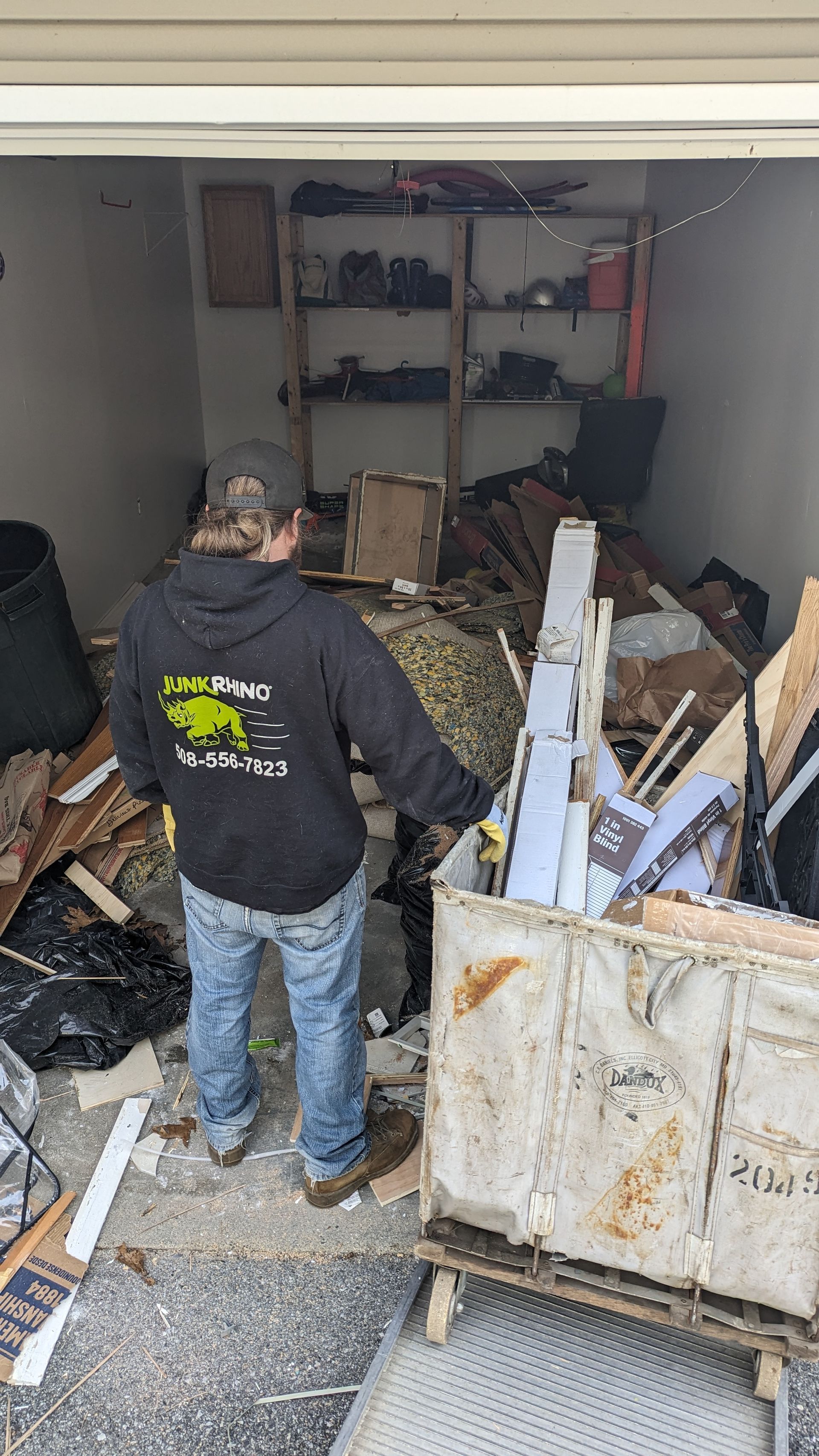 Man in a hoodie stands in a messy garage, looking at debris in a metal container, shelves in the background.