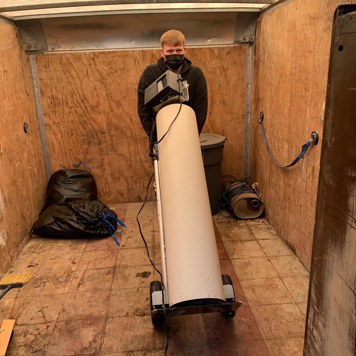Man in a mask holds a tall, white machine inside a wooden cargo area. Black trash bag and equipment are nearby.