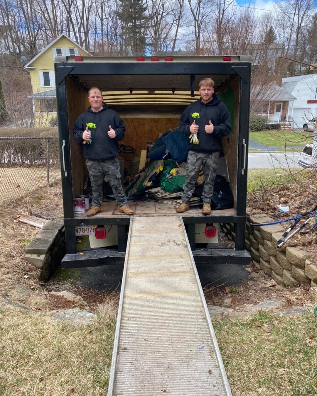 Two people stand in a moving truck with thumbs up, holding screwdrivers. Ramp extends from the truck.