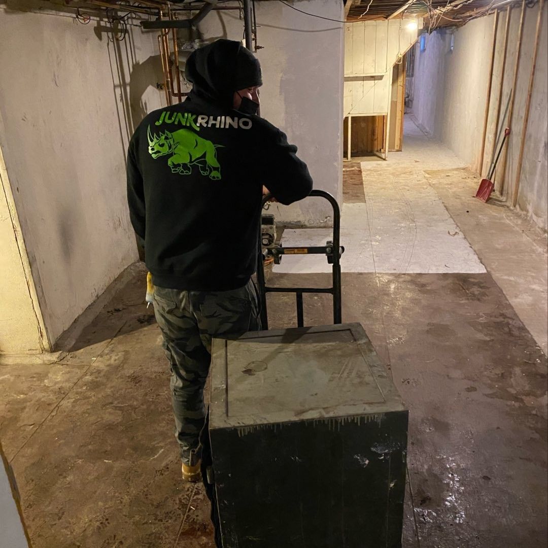Man from Junk Rhino wearing a black sweatshirt and hat moves a safe with a dolly in a basement.