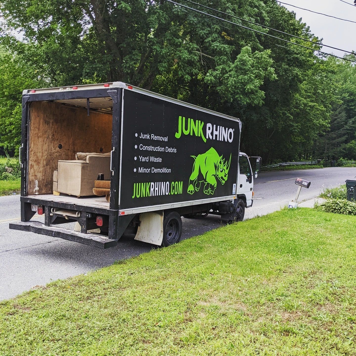 Junk Rhino truck parked on a residential street, with open back containing furniture. Green grass and trees visible.