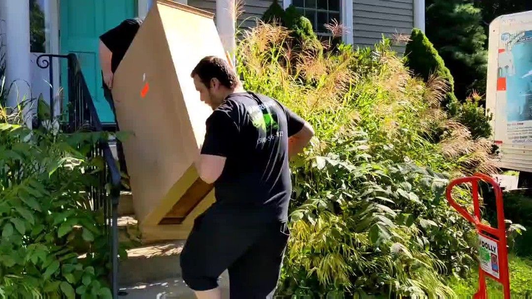 Two people carrying large cardboard box up outdoor steps to a house.