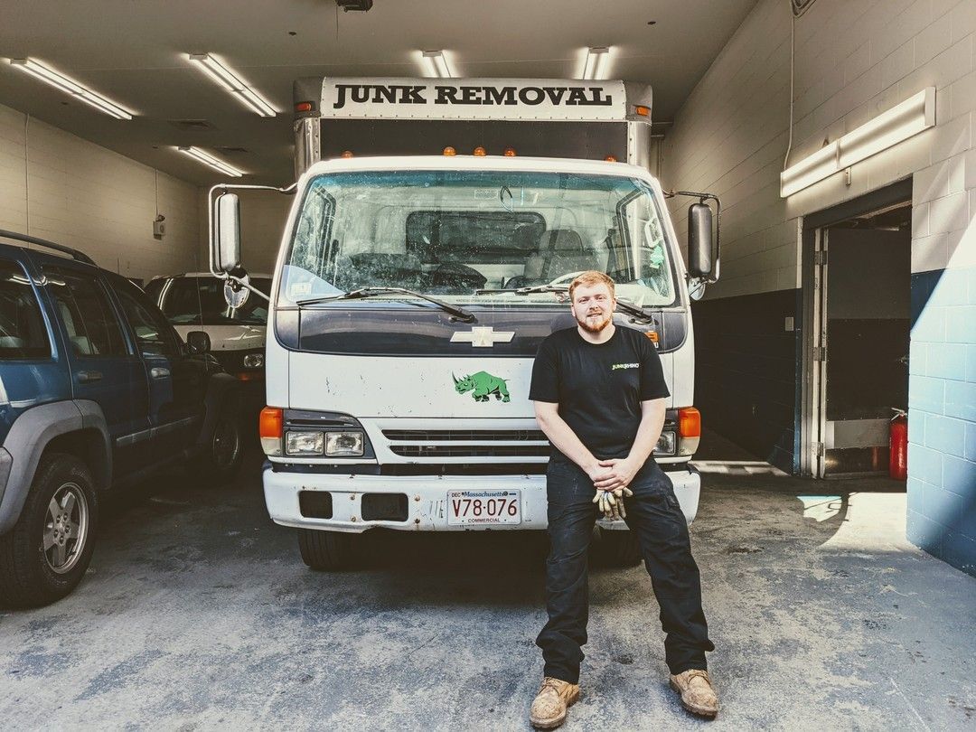 Man in black work clothes sits in front of a junk removal truck. The truck is white with 