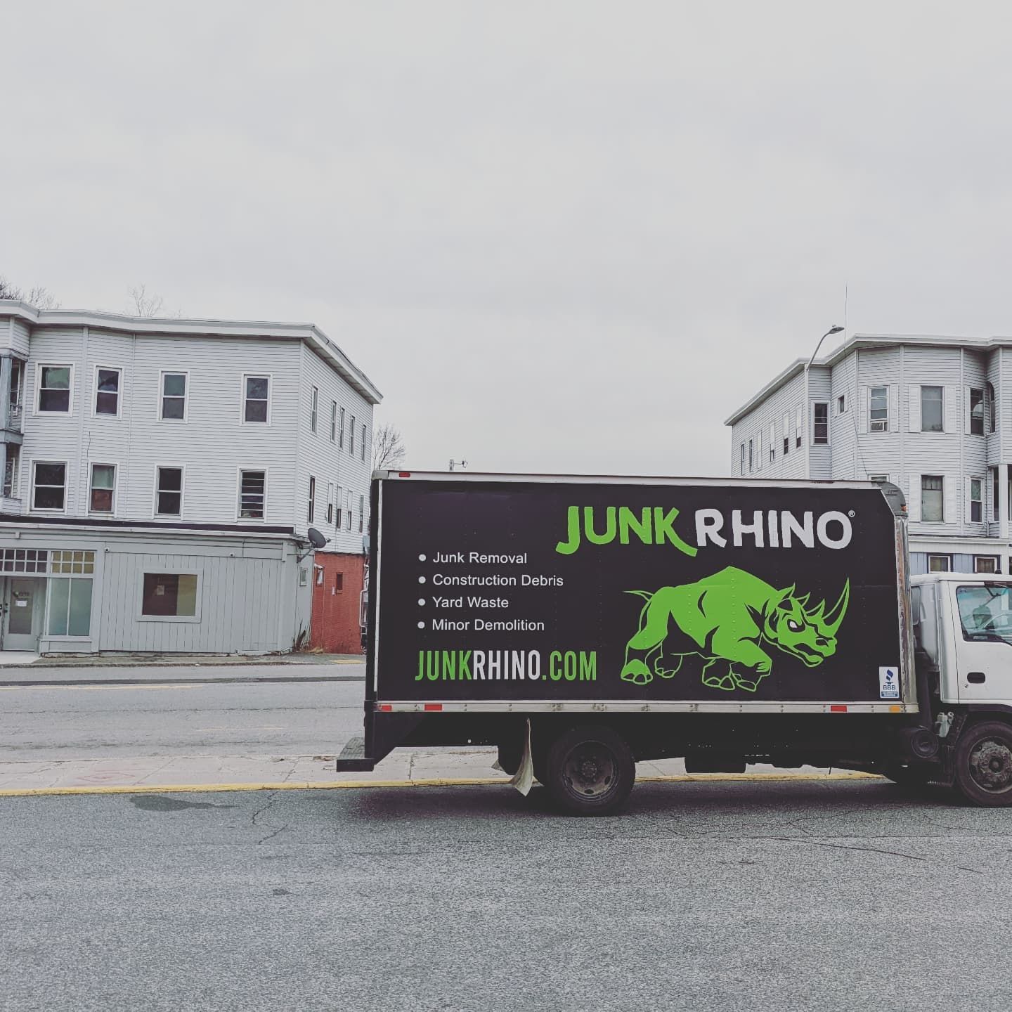Junk Rhino truck parked in front of white buildings with boarded-up windows on a cloudy day.
