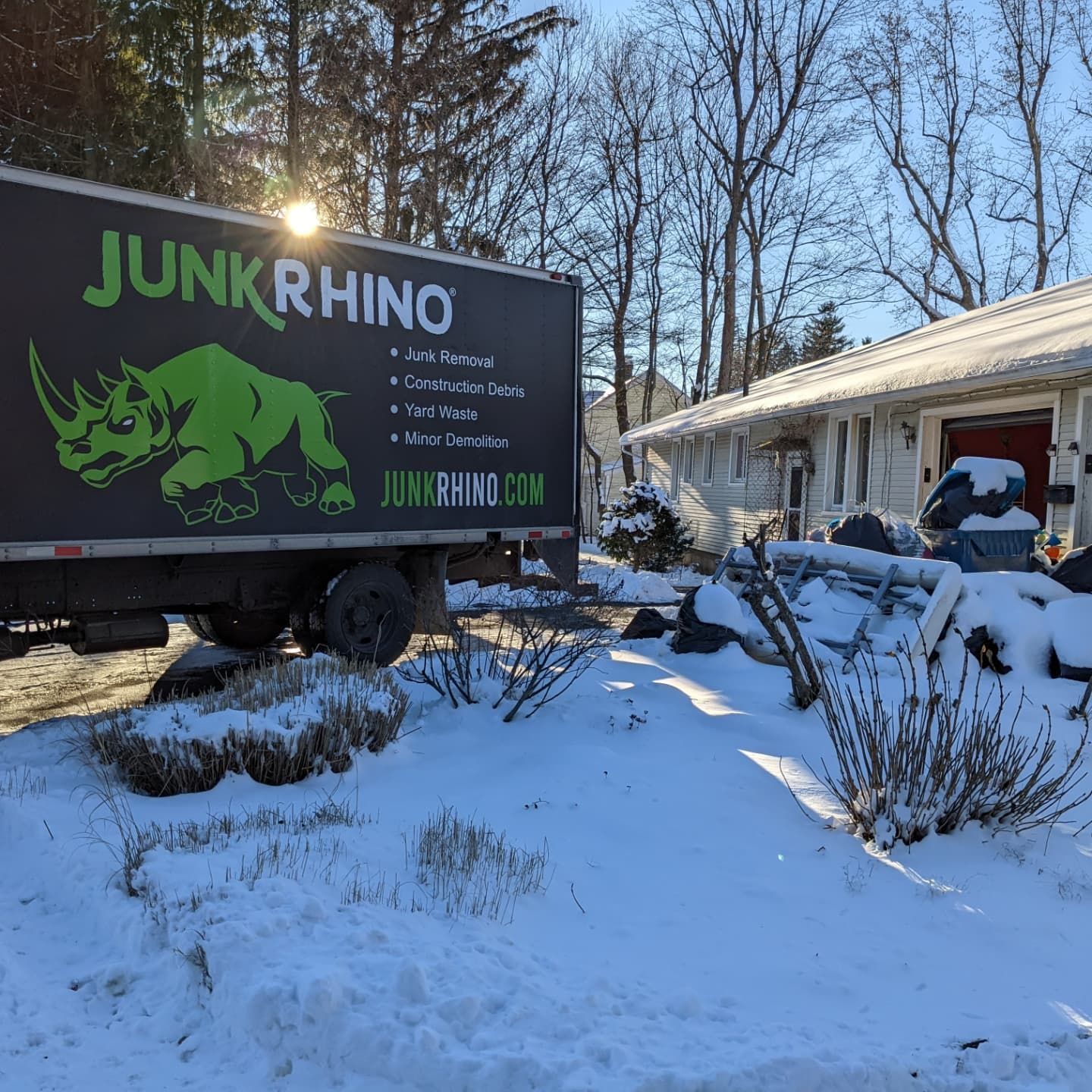 Junk Rhino truck parked outside a snow-covered house; sun shining.