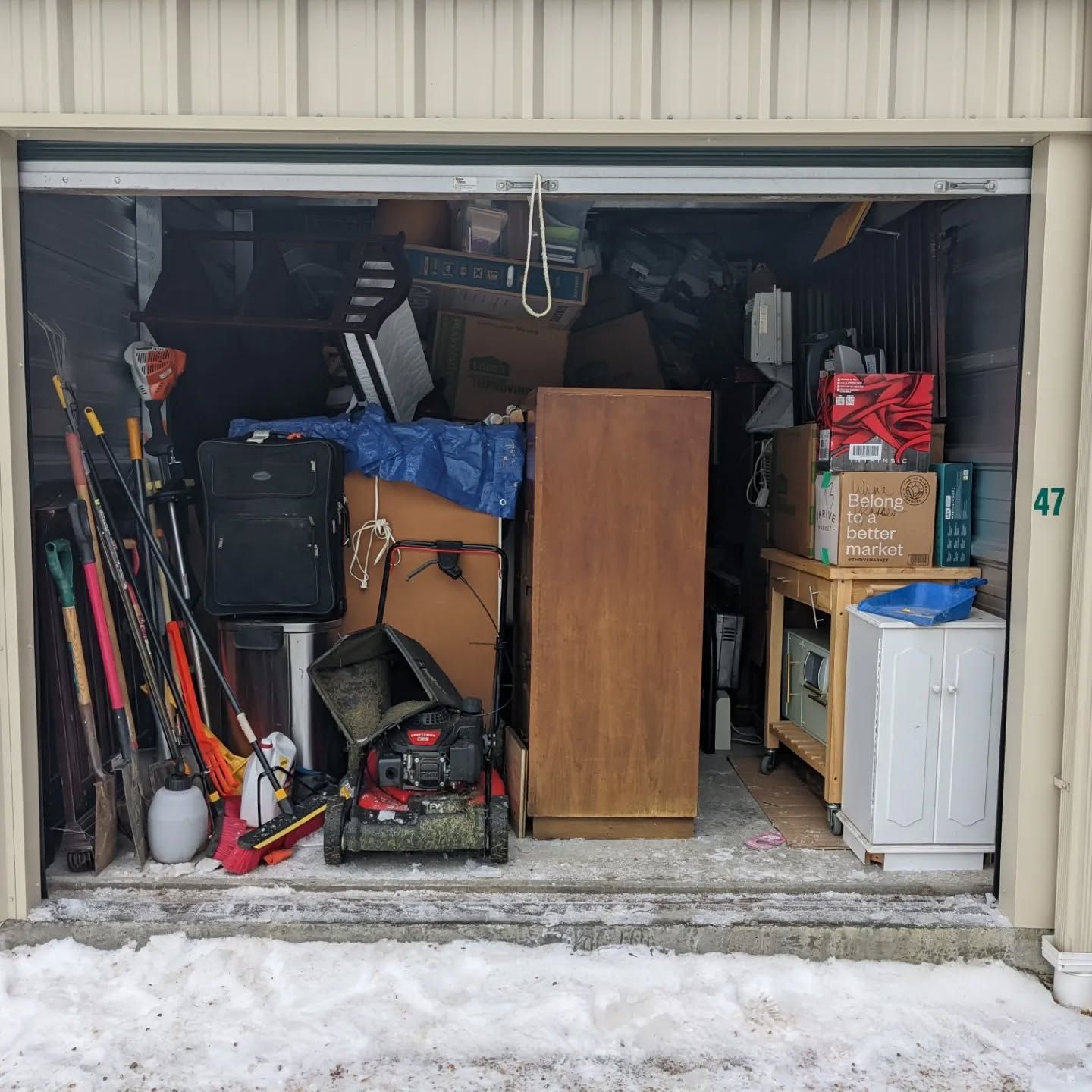 Storage unit filled with packed boxes, luggage, tools, and furniture. Snowy ground outside.