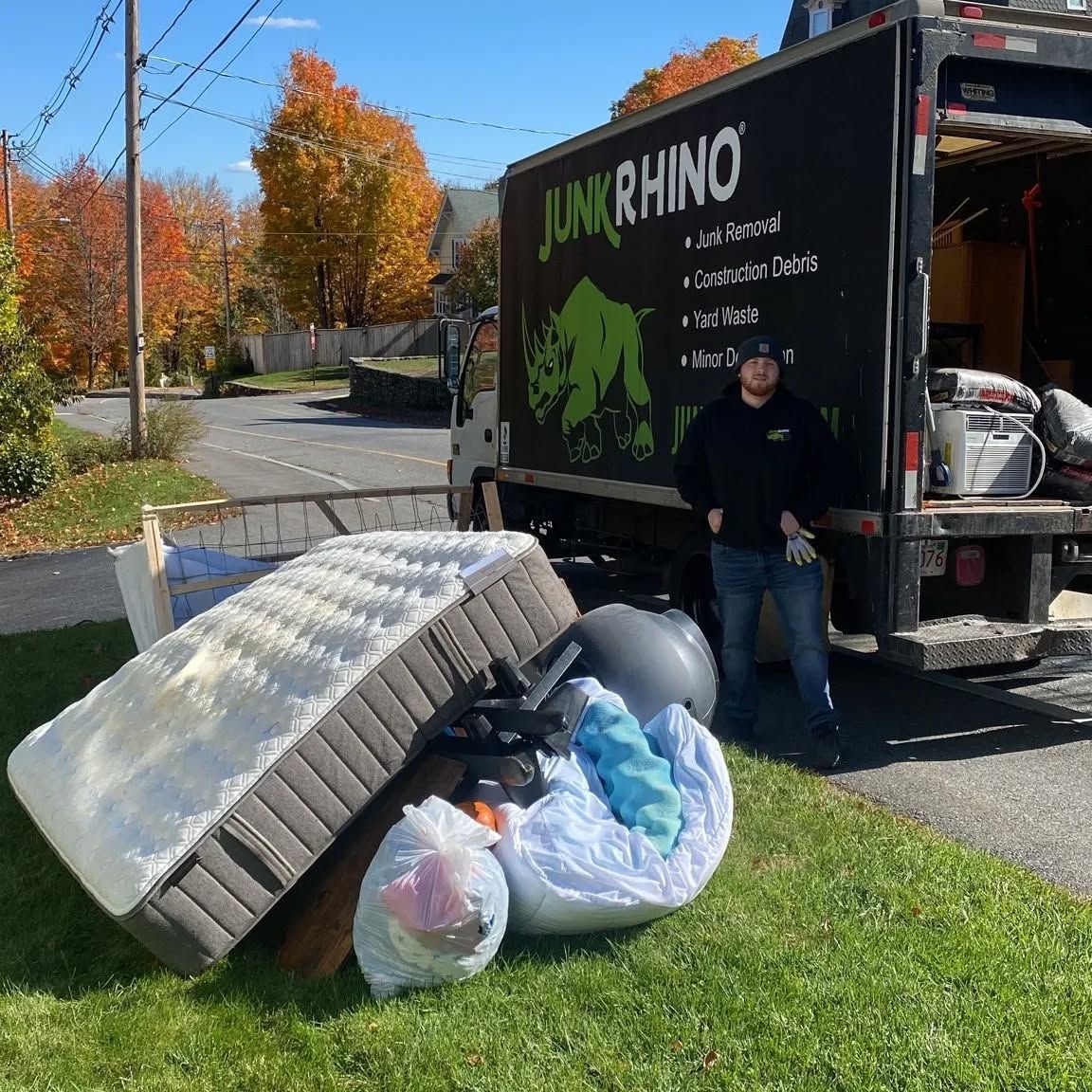 A Junk Rhino truck with a person standing next to discarded items, including a mattress and bags, on grass.