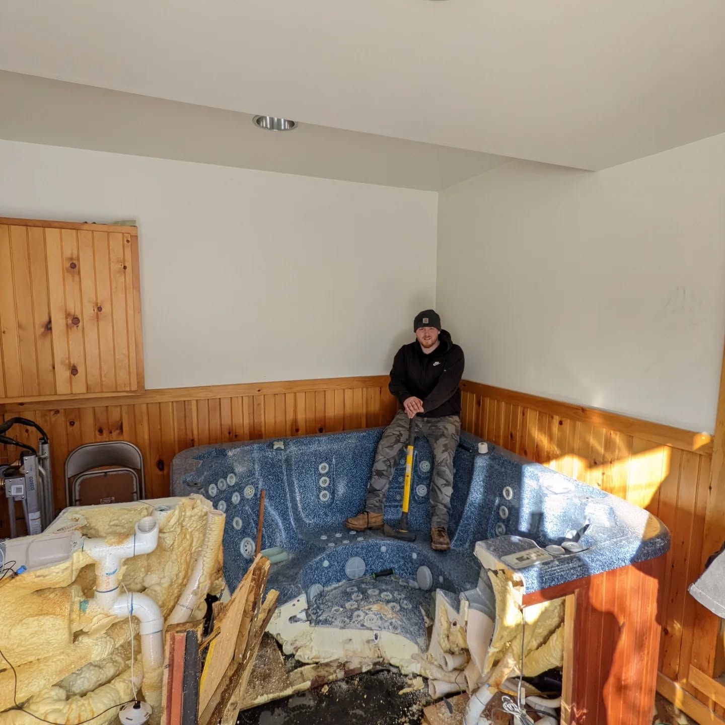 Man sits on a damaged blue hot tub indoors, surrounded by debris.