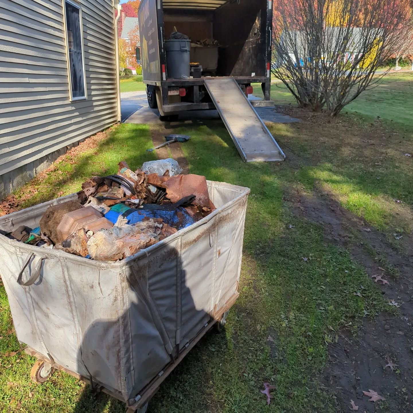 A large bin filled with debris being loaded into a truck with a ramp in a yard.