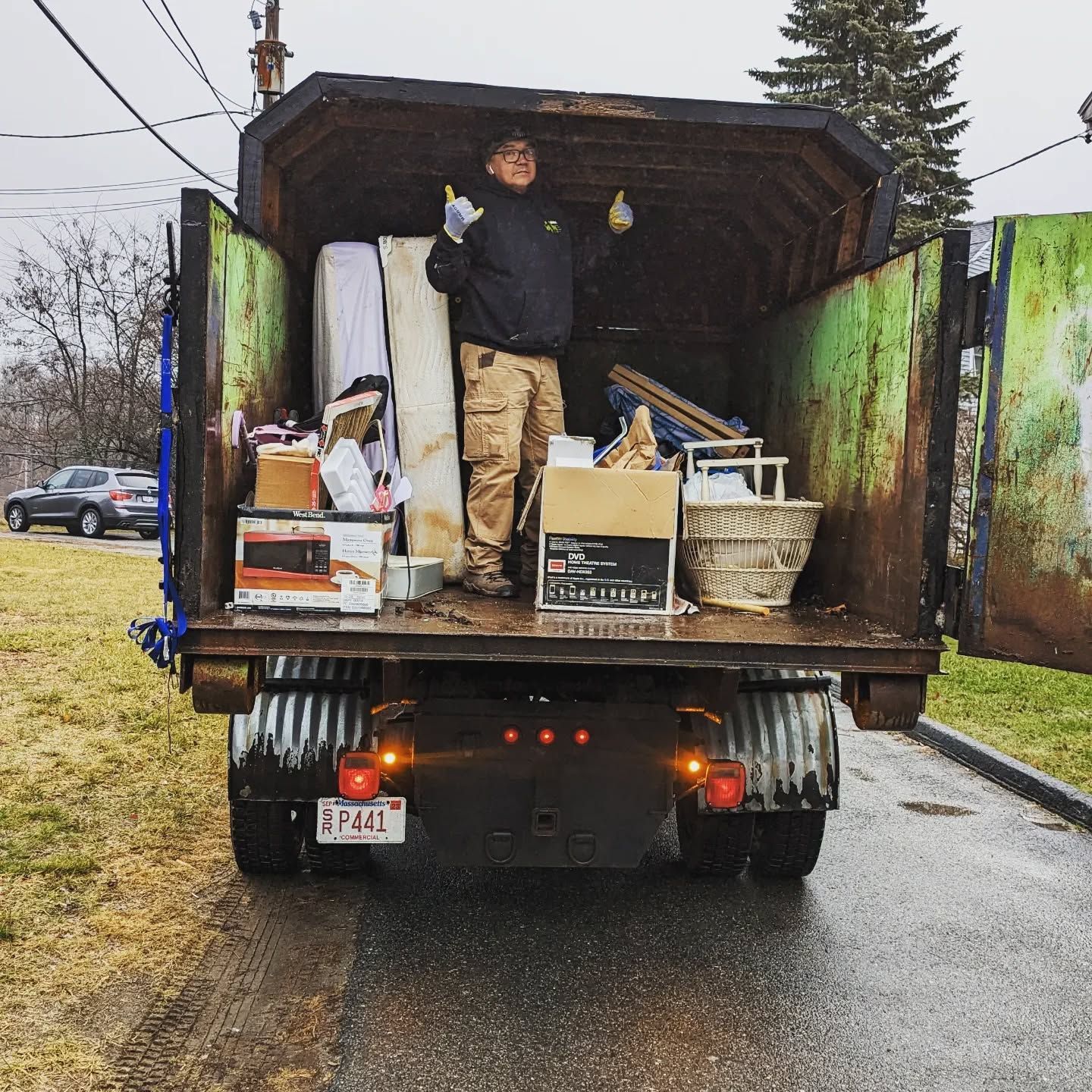 Man in a garbage truck with debris, giving a thumbs up. Exterior shot, cloudy day.