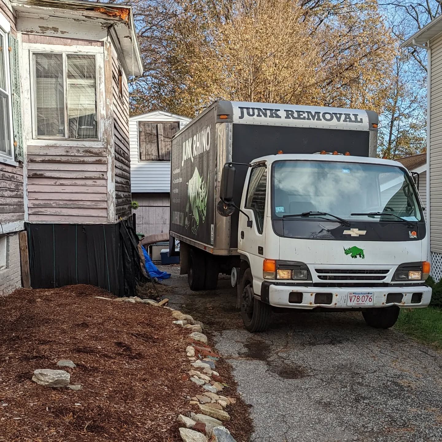 Junk removal truck parked beside a weathered house, ready to load debris. Brown mulch lines the yard.