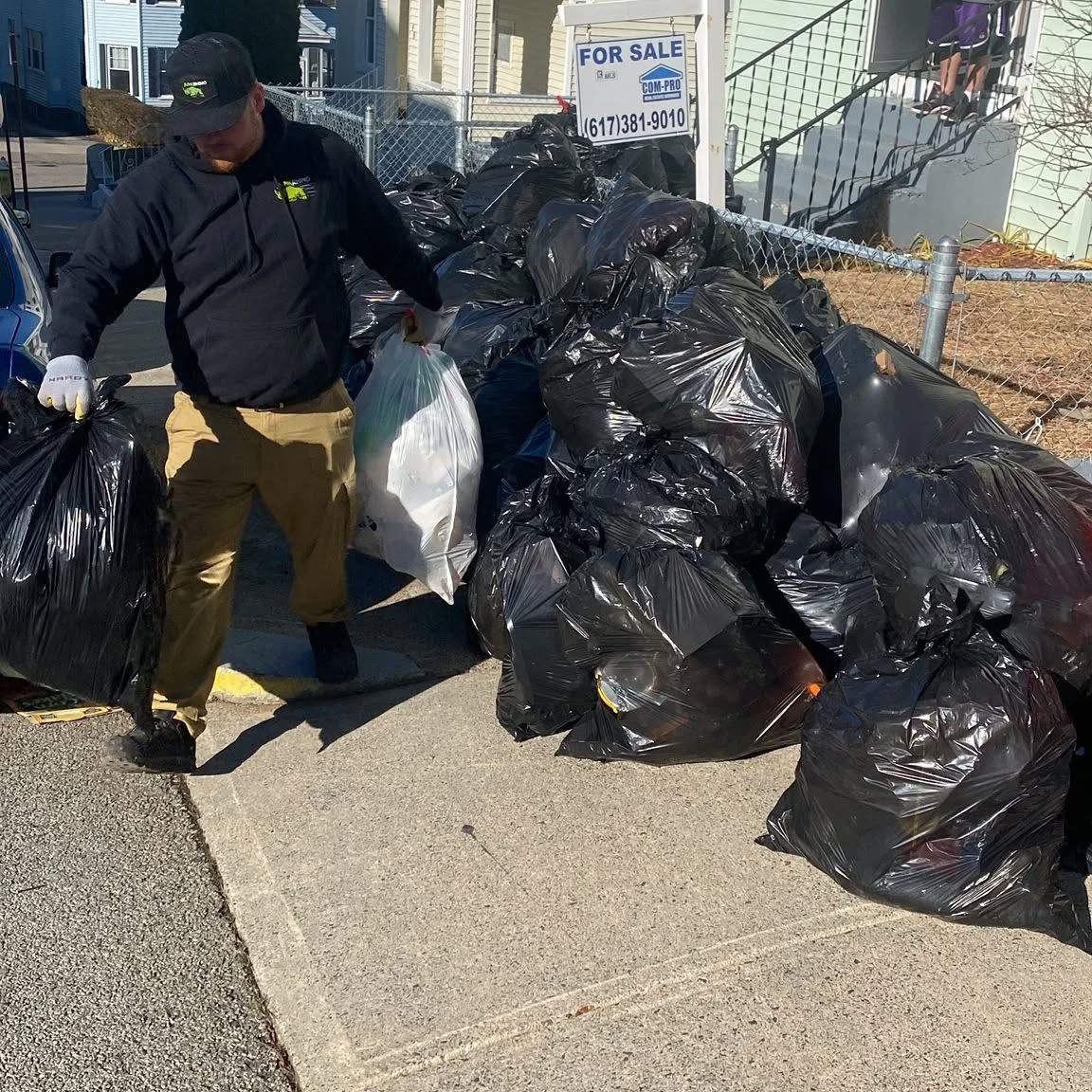 Man carries bags of trash near a pile of trash bags on a sidewalk, next to a house with a 