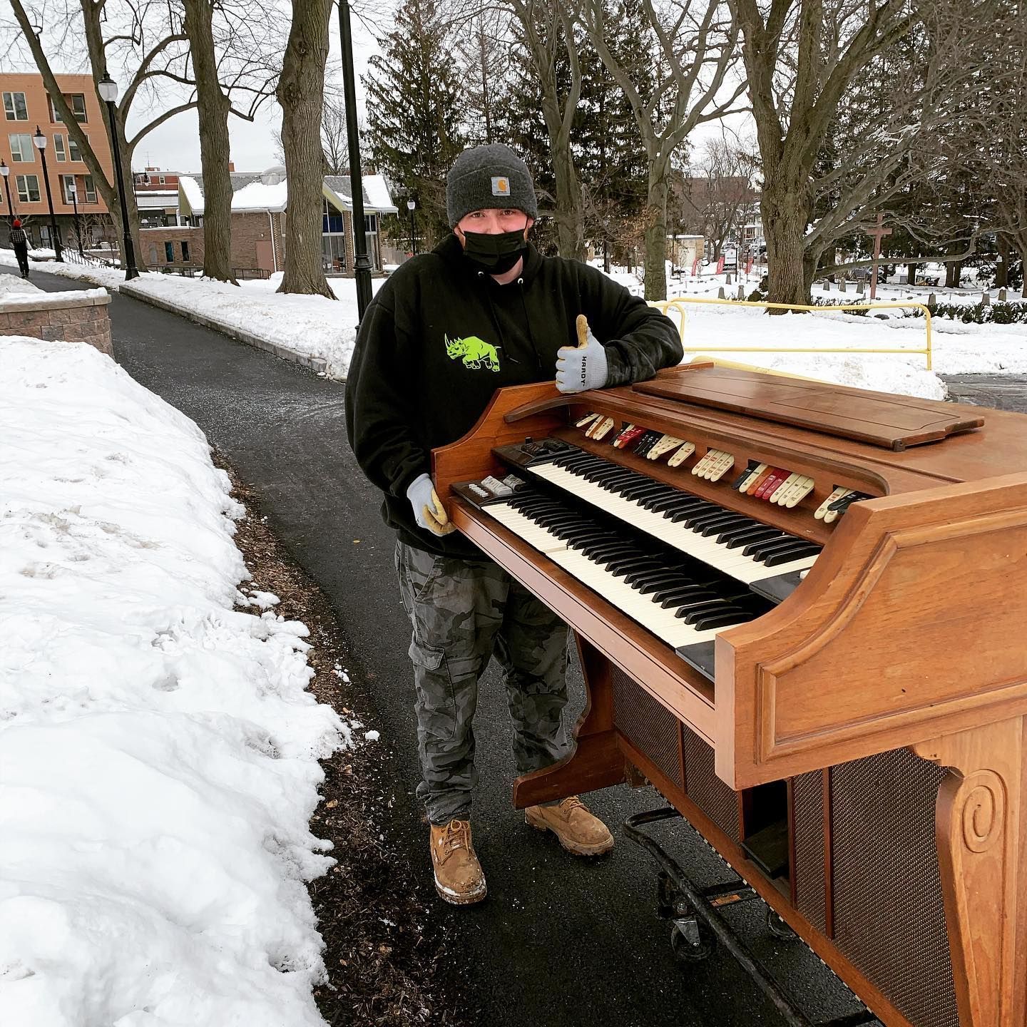 Person in black shirt, cap, and mask giving thumbs-up next to an organ on a snowy sidewalk.