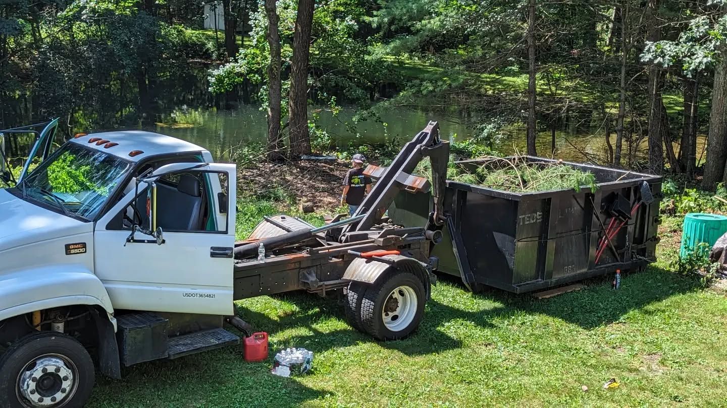 White truck with a dumpster in a grassy area, trees in the background. Dumpster is filled with yard waste.
