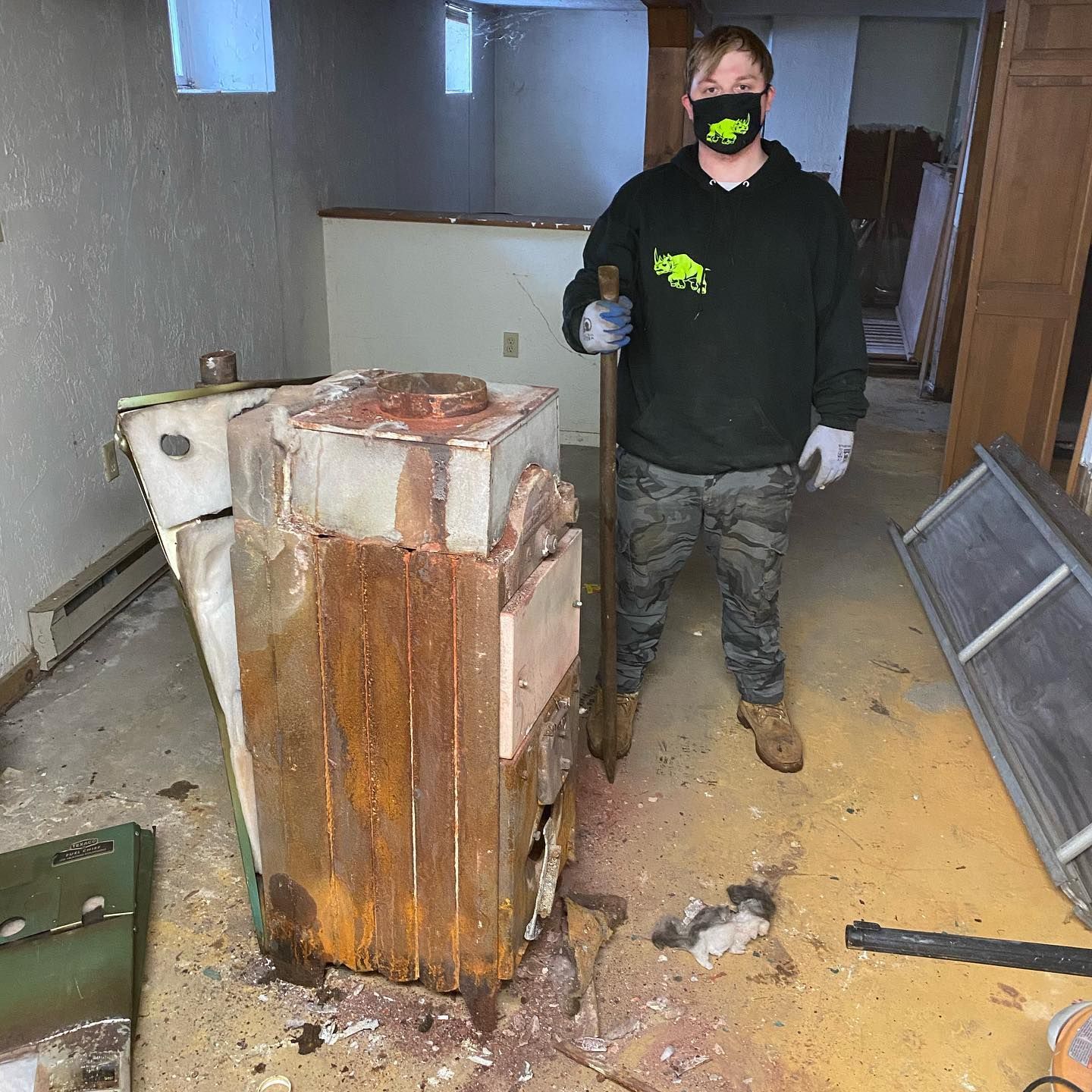 Man standing near a damaged heater in a basement, holding a tool. He wears a mask and work clothes.