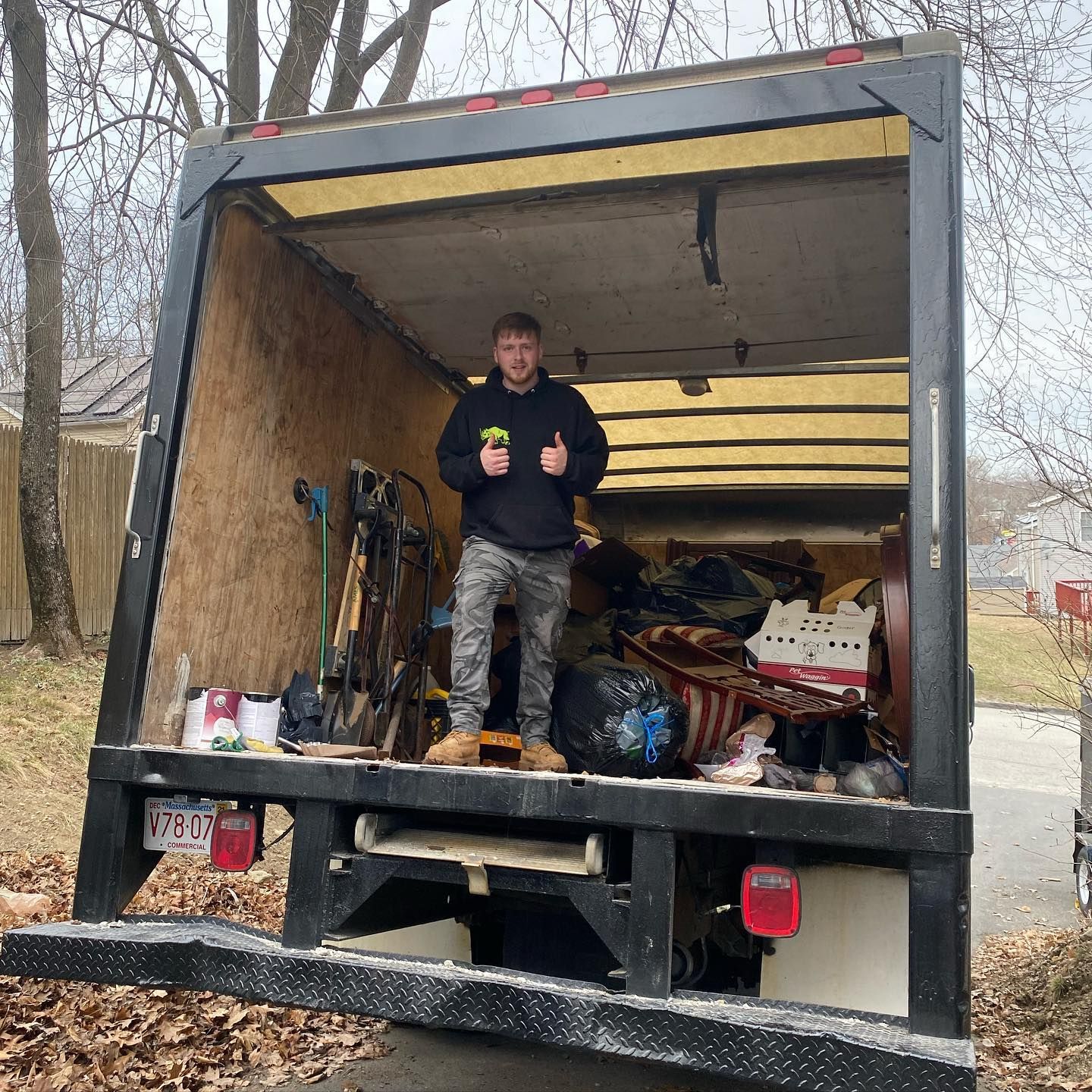 Man standing in the back of a moving truck with thumbs up, surrounded by items. Outdoor setting.