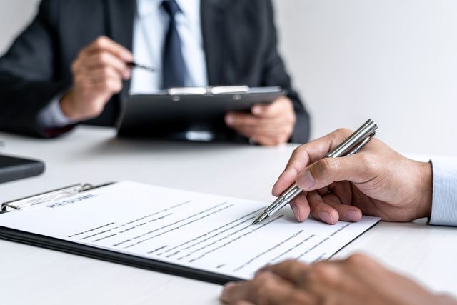 Person signing a document with another person observing at a desk.