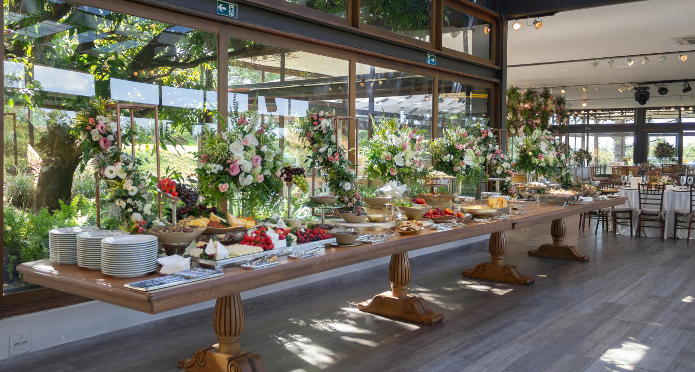 A long wooden banquet table laden with food and floral arrangements in a bright, modern room with floor-to-ceiling windows.