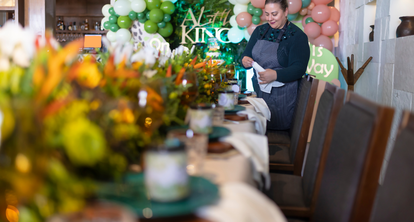 A person prepares a long dining table for an event, adorned with vibrant floral arrangements and festive decorations.
