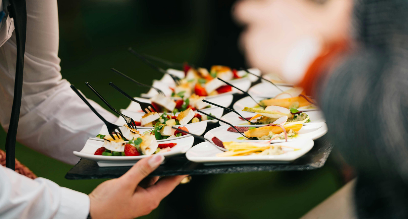 A server holds a tray of small white plates featuring appetizers with fruit, cheese, and dark picks at a catered event.