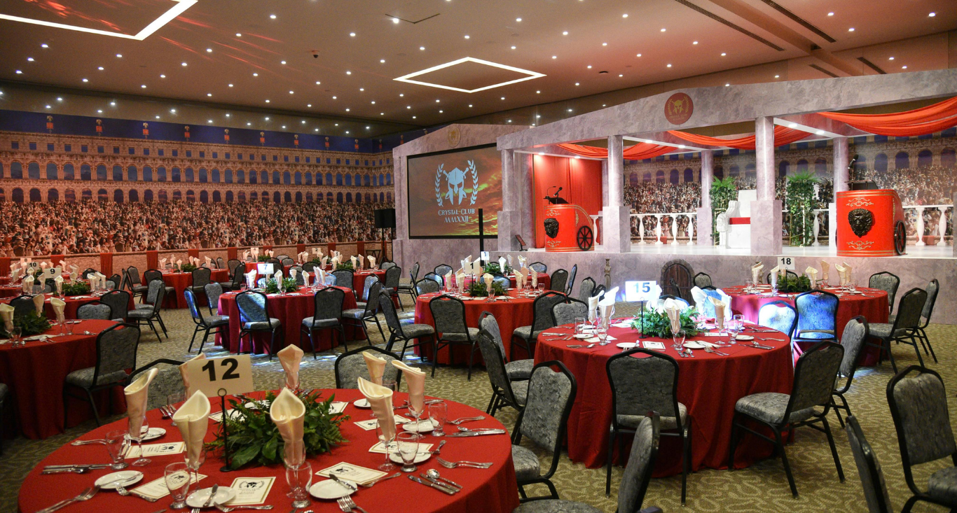 A decorated banquet hall with red tablecloths, table number 12, and a stage with classical columns and a Roman mural.