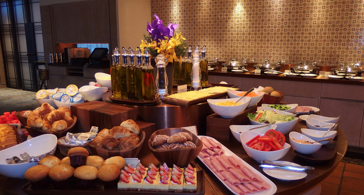 A hotel breakfast buffet featuring a variety of pastries, fresh fruit, sliced meats, vegetables, and condiments.