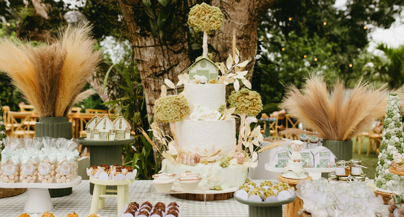 A three-tiered white wedding cake surrounded by treats, pampas grass, and floral decorations on an outdoor table.