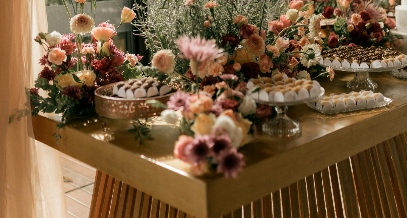 A wooden table decorated with vibrant floral arrangements and platters of assorted small desserts.