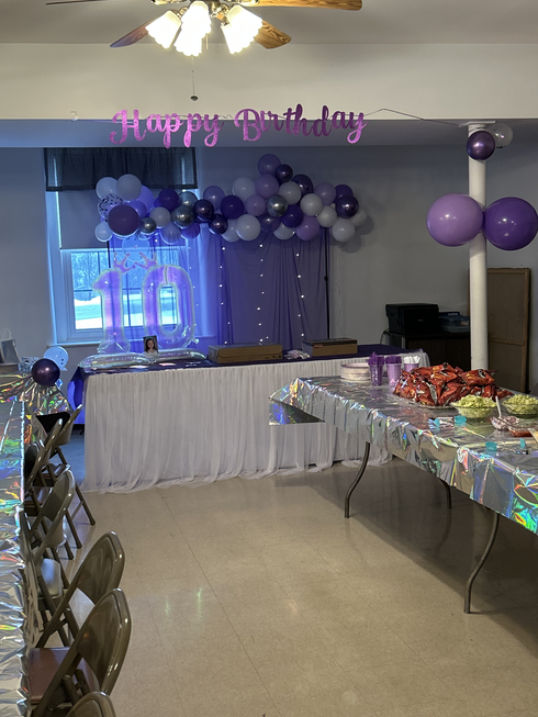 A party space decorated for a 10th birthday with a purple, white, and silver balloon arch, streamers, and tables.