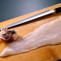 Un filete de pescado blanco crudo con un cuchillo japonés sobre una tabla de cortar de madera.