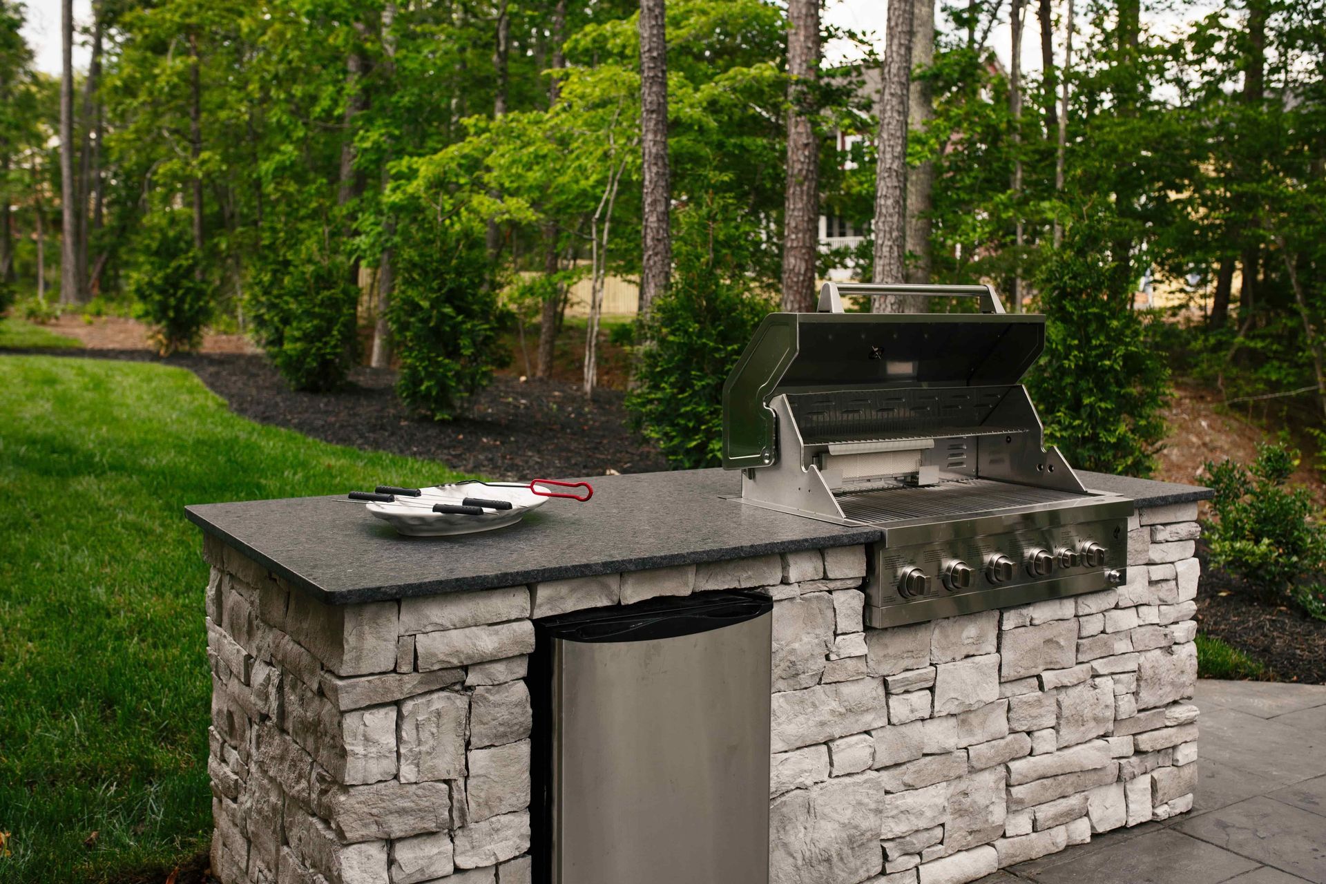 Outdoor kitchen with stone facade, grill, countertop, and refrigerator, surrounded by greenery.