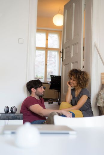 A man and a woman are sitting on the floor in a living room talking to each other.