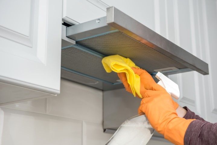 A worker in an orange glove, yellow towel, and a sprayer is cleaning the kitchen hood.