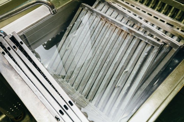 Metal kitchen exhaust filters soaked in a commercial stainless steel sink filled with water for cleaning and degreasing.