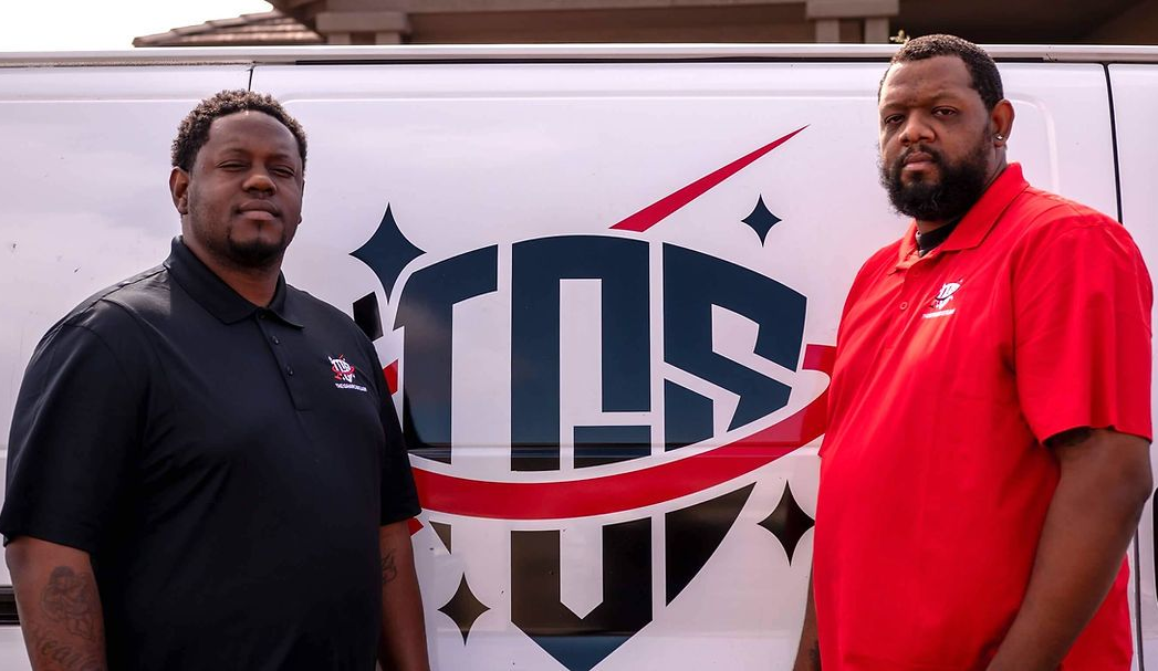 Two men stand in front of a van. The van has a logo that reads