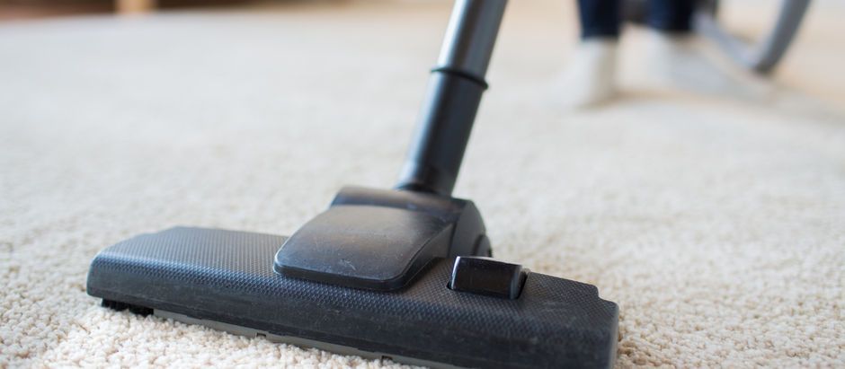 Vacuum cleaner on beige carpet, cleaning floor. Person's legs visible in background.