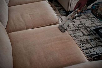 A person vacuuming a light-colored sofa with a handheld upholstery cleaner.