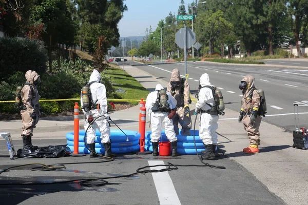 Squadra Hazmat in tute protettive decontamina attrezzature vicino a una strada; piscina blu, alberi, giornata di sole.