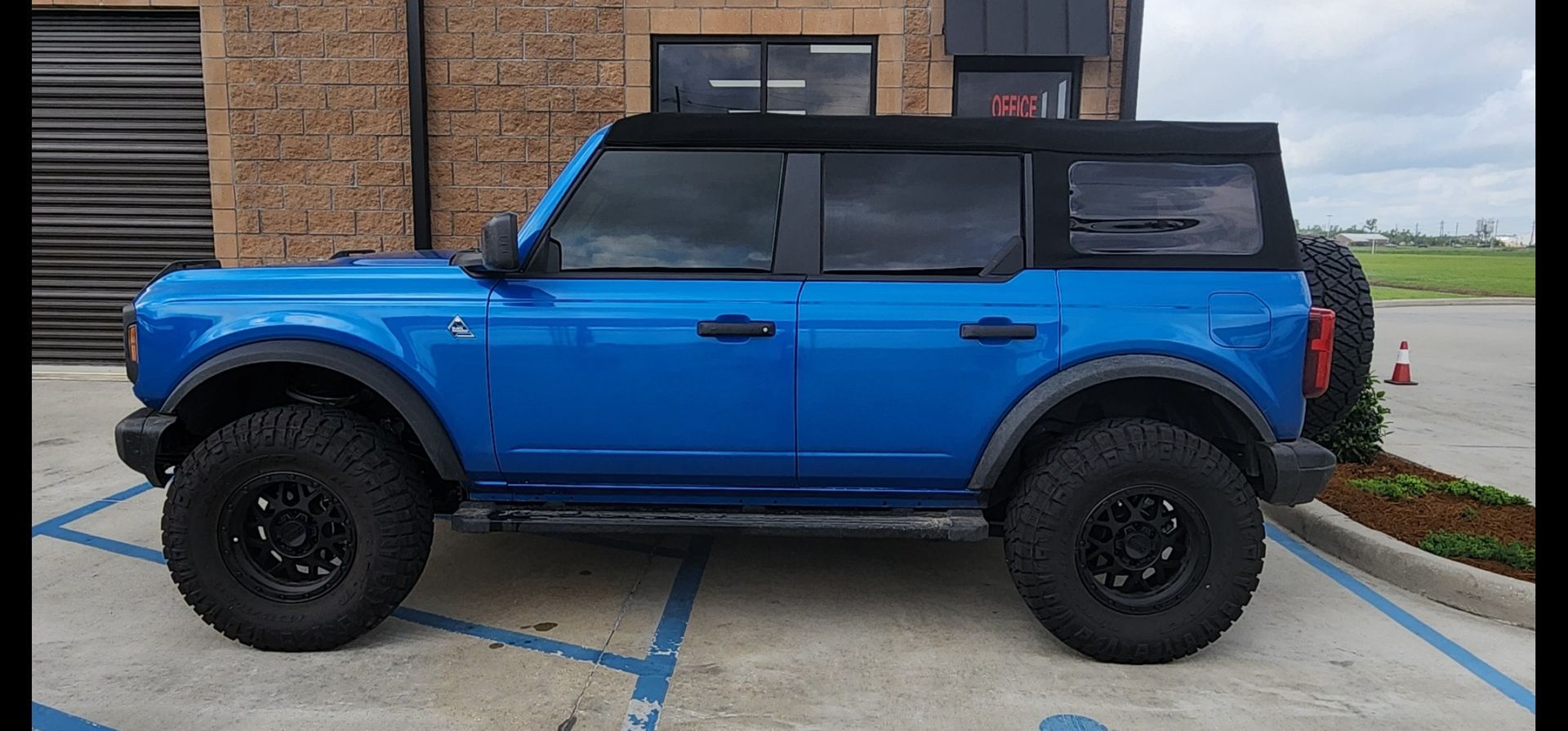 A blue 4-door Ford Bronco with a black soft top and rugged off-road tires parked in a paved lot.