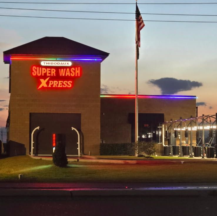 A white car being cleaned in an automated car wash, covered in soap suds with blue rotating brushes on the sides.