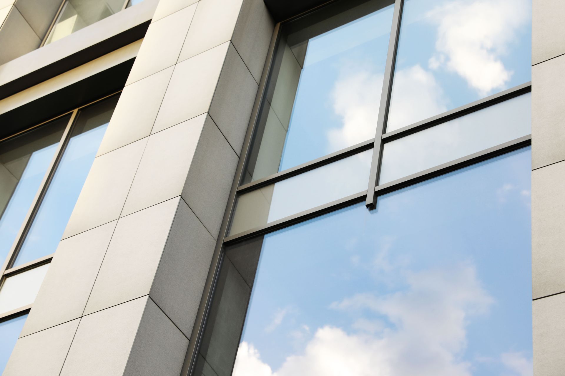 Modern building facade with light-colored stone panels and large glass windows reflecting a cloudy blue sky.