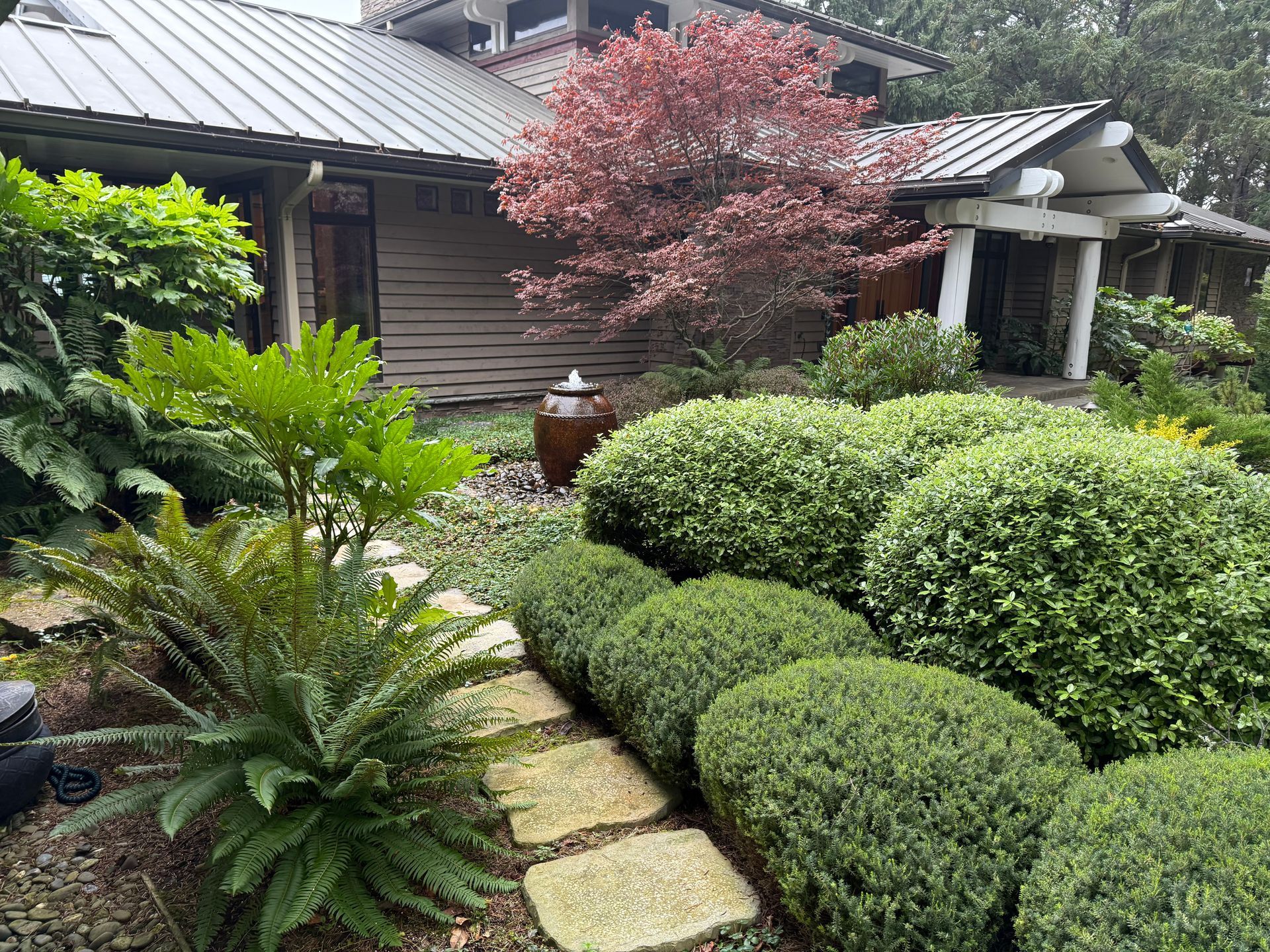 Curving paved path through a vibrant garden with colorful azaleas, trimmed trees, and lush green grass under a clear blue sky.