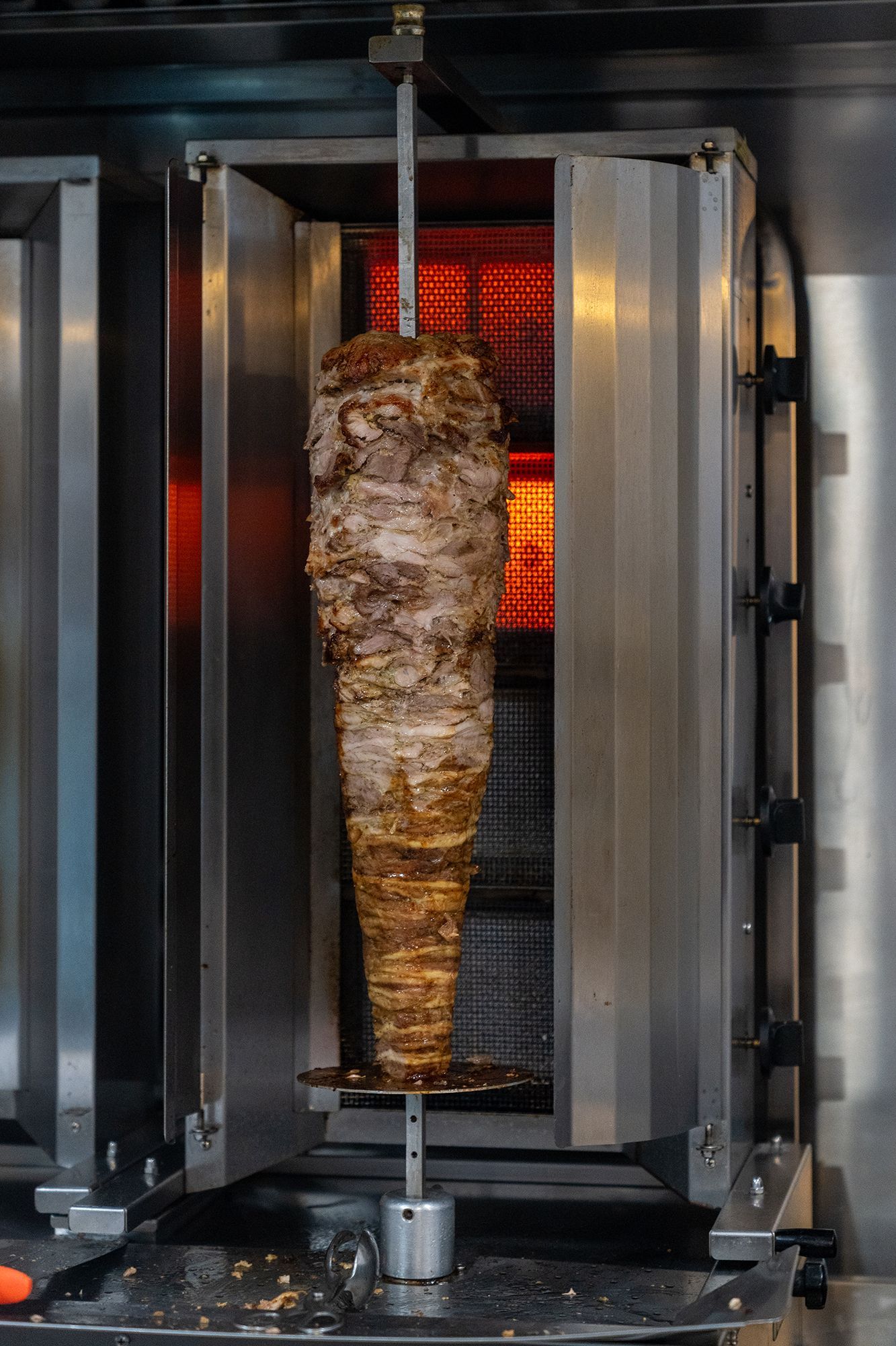 Spiedo verticale di carne condita che ruota in un girarrosto in acciaio inox, con una fonte di calore rosso acceso sullo sfondo.
