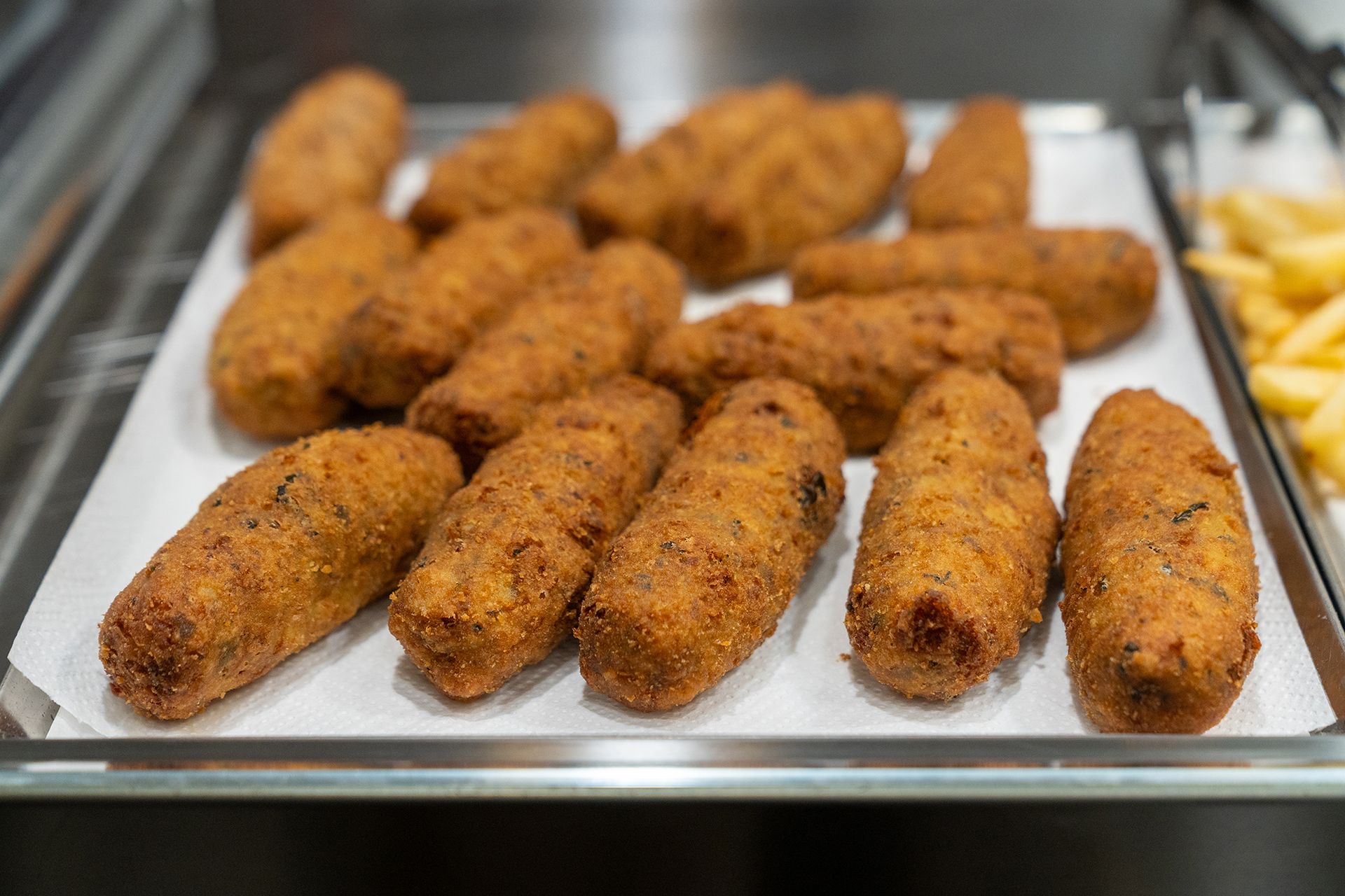 Vassoio di crocchette impanate e dorate su carta bianca, accanto a un vassoio di patatine fritte.