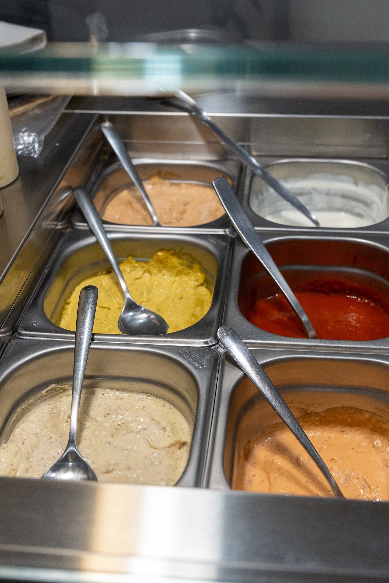 Contenitori di metallo riempiti con varie salse; in ognuno di essi sono appoggiati i cucchiai.