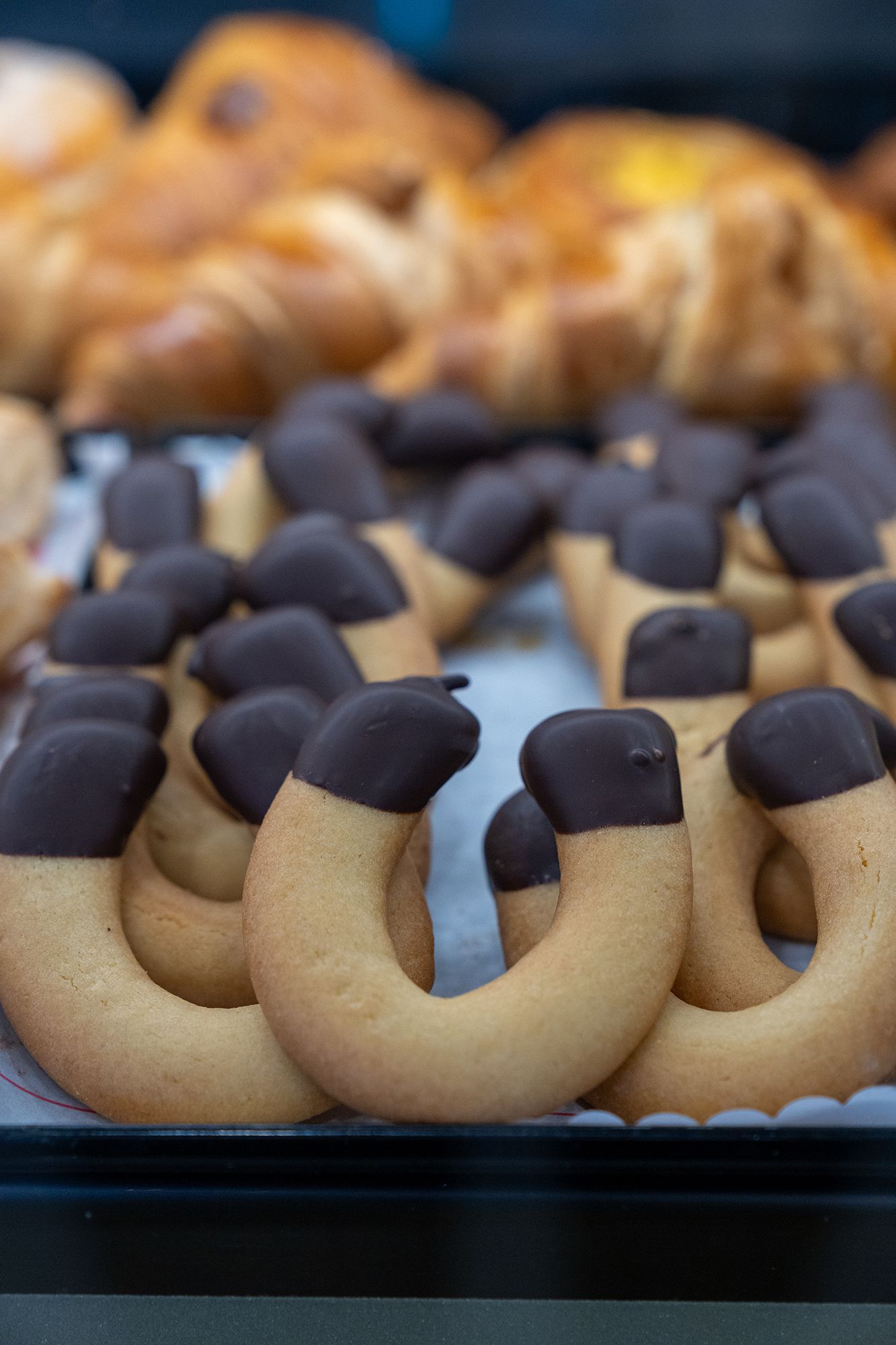 Biscotti a forma di U immersi nel cioccolato fondente, vassoio di pasticcini sullo sfondo.
